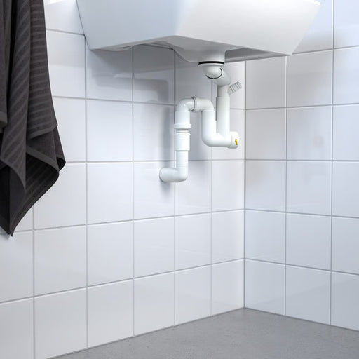 The IKEA RÄNNILEN flexible white water trap installed underneath a white bathroom sink, showing its pipe connecting to a wall drain against white tiled walls-30562433