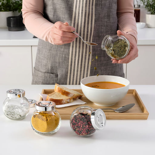 Person seasoning soup using IKEA RAJTAN glass spice jar with bread and bowl on wooden tray