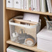 Storage basket with handles on a wooden shelf with books and office supplies.