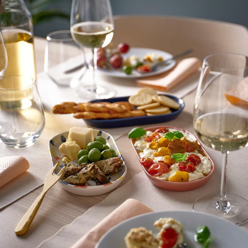 A dining table set with white wine glasses and various appetizers served on IKEA RÖDSNULTRA oval plates: one pink with burrata and tomatoes, one blue with crackers and breadsticks, and one white with colorful splatters holding olives and cheese.60604506