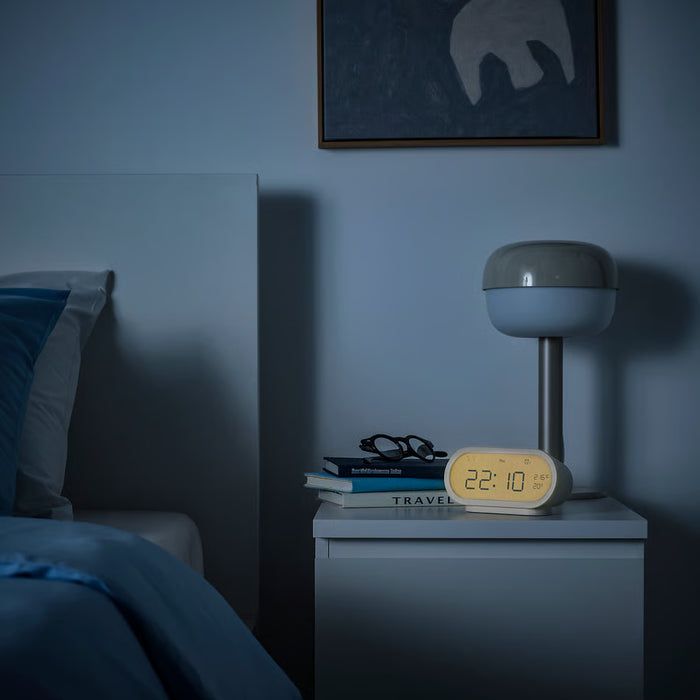 Bedroom nightstand with lamp, books, and alarm clock against a dark blue wall.