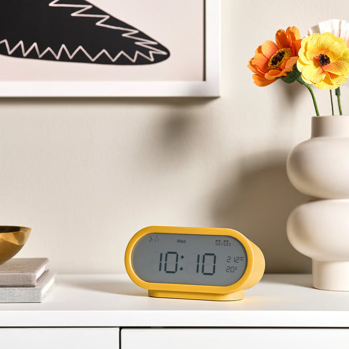 Yellow alarm clock displayed on a shelf with books, vase and artwork