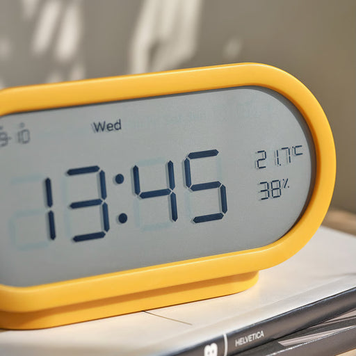 Yellow digital alarm clock displaying time, temperature and humidity on a bedside table