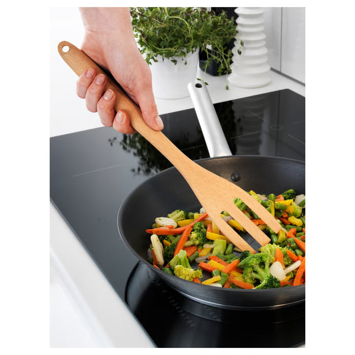 Hand using IKEA RÖRT wooden fork to stir vegetables in a black non-stick pan on a stove
