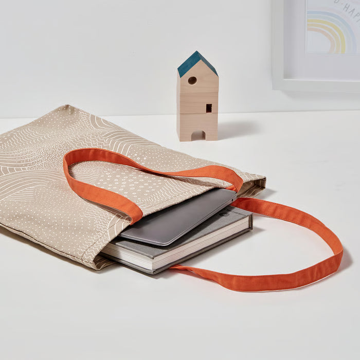 Beige tote bag with orange strap holding books on a white surface with a small wooden house model.