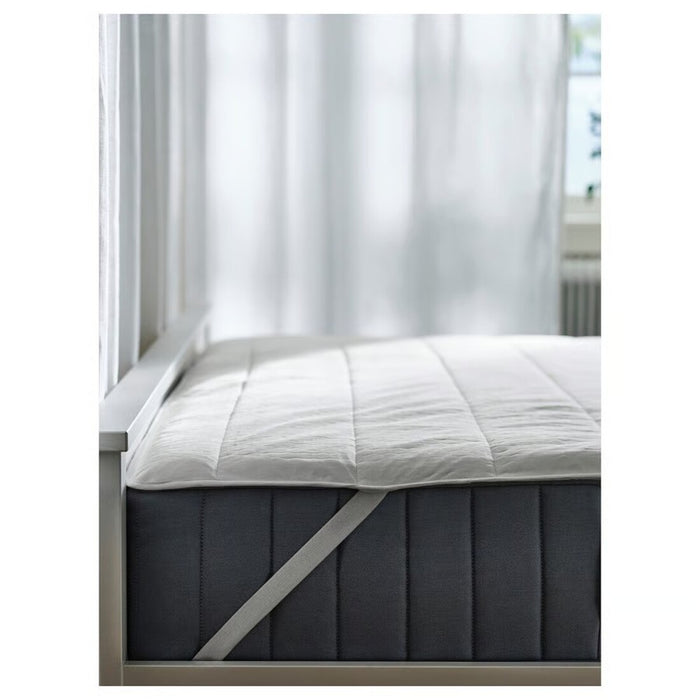 Gray mattress with white cover on a bed frame, against a white curtain background