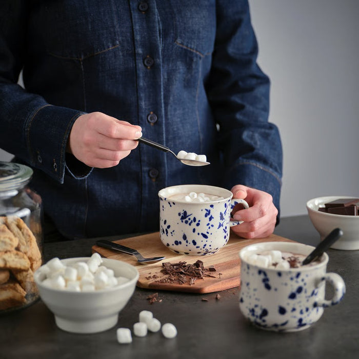 "A breakfast setup on a wooden table featuring a glass pitcher of milk, a jar of granola, a light green bowl, plates with a croissant and a slice of cheese, sliced bread on a wooden cutting board, a fork, and a striped cloth napkin, illuminated by soft natural light.