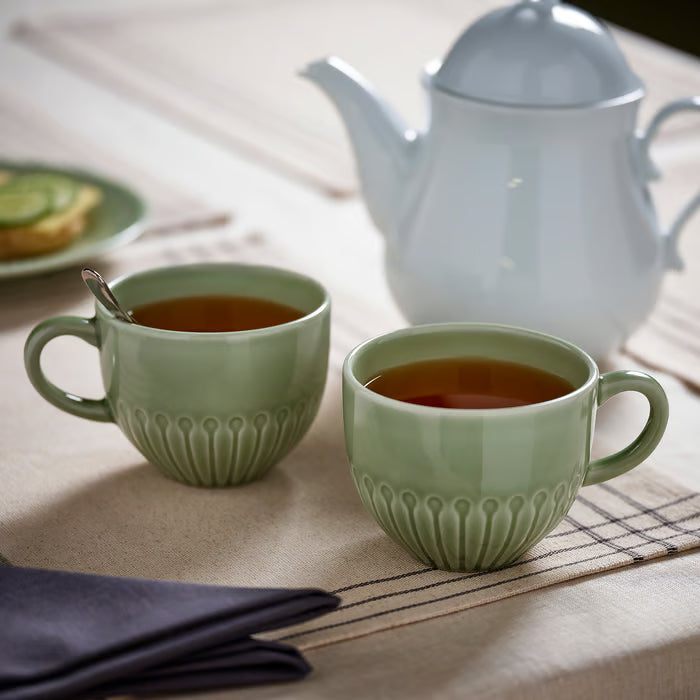 Two IKEA STRIMMIG pale grey-green mugs filled with hot drink on table with white teapot and linen napkins