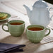 Two IKEA STRIMMIG pale grey-green mugs filled with hot drink on table with white teapot and linen napkins