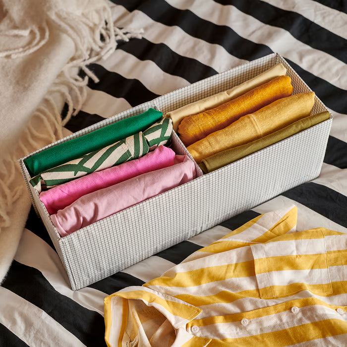 Two IKEA STUK white black grid storage boxes holding folded colourful fabrics on striped bedspread