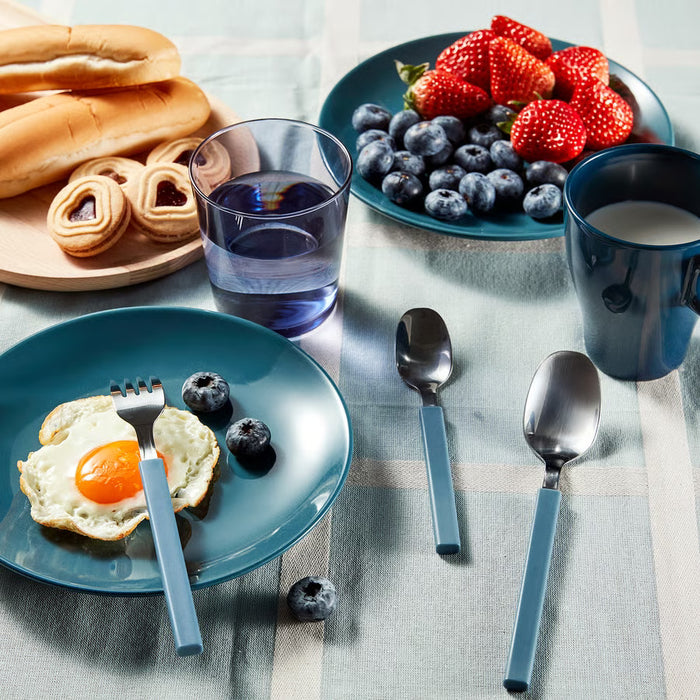 A styled breakfast scene on a tablecloth with blue plates holding a fried egg with a blue-handled fork, blueberries, strawberries, bread, a blue drink, a blue mug, and two blue-handled metal spoons.