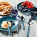 A styled breakfast scene on a tablecloth with blue plates holding a fried egg with a blue-handled fork, blueberries, strawberries, bread, a blue drink, a blue mug, and two blue-handled metal spoons.