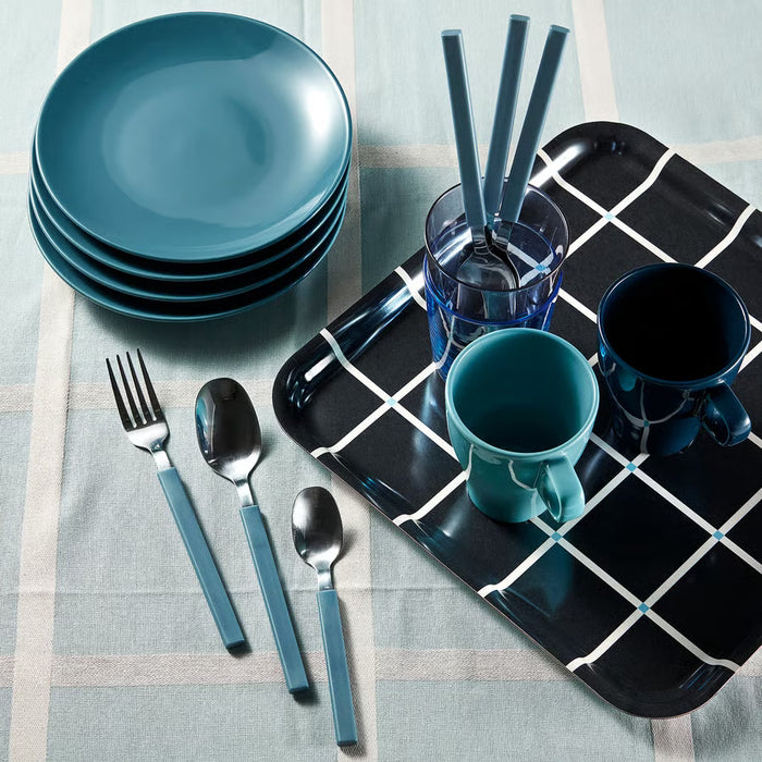 A place setting with teal and navy blue ceramic tableware, including plates, mugs, cups, spoons, and forks, arranged on a plaid tablecloth alongside a black tray with a white grid pattern.