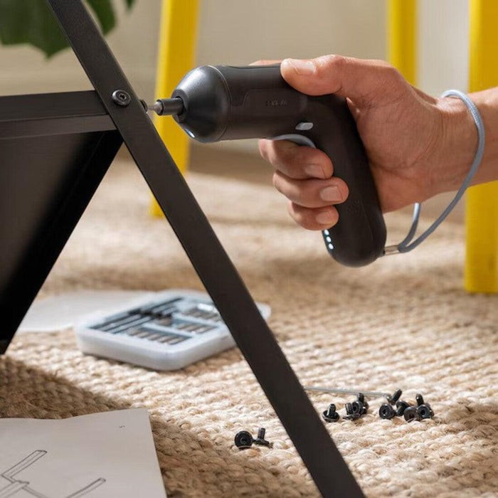 A person is using a black electric screwdriver to assemble a piece of furniture. The screwdriver is being used to tighten a screw on a metal frame. There are several screws scattered on a carpeted floor, along with an open toolkit and an instruction manual. The scene suggests a DIY furniture assembly process- 