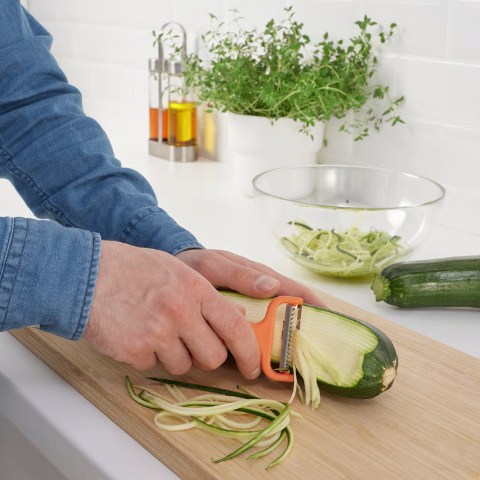 Person peeling courgette into long strips with orange IKEA UPPFYLLD cutter on wooden board, bowl of strips in background