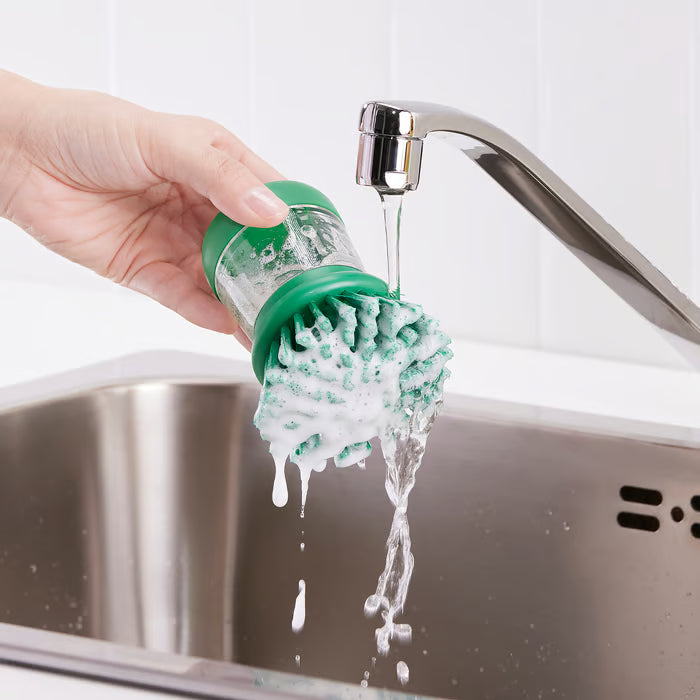 Person holding IKEA VIDEVECKMAL green dish brush under tap to rinse while dispensing soap