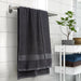 IKEA VITBACKEN dark grey bath towel hanging on a silver rail, larger than folded towels on a white shelf with a plant