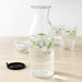 Digital Shoppy Ikea carafe with lid , An image of an IKEA carafe with a clear glass body and a lid made of cork. The carafe is filled with ice water and placed on a blue picnic blanket in a park. 302.919.22