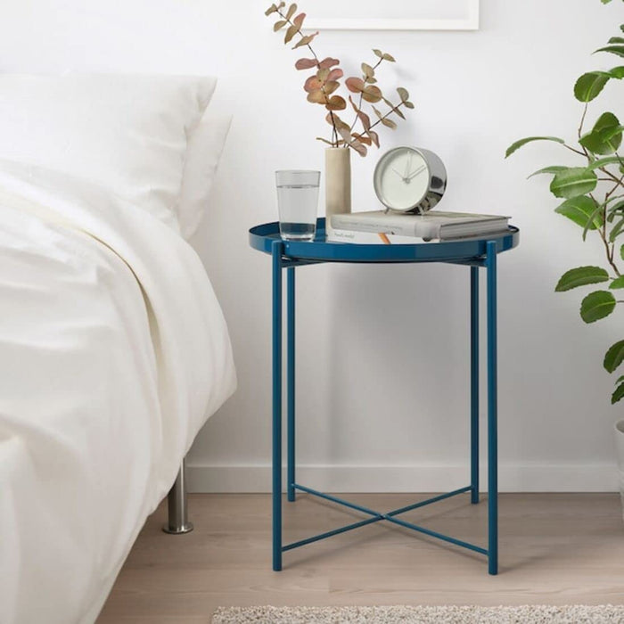 "The IKEA tray table in pale shown in a bedroom setting. It is being used as a nightstand, highlighting its functional design and versatility in small space