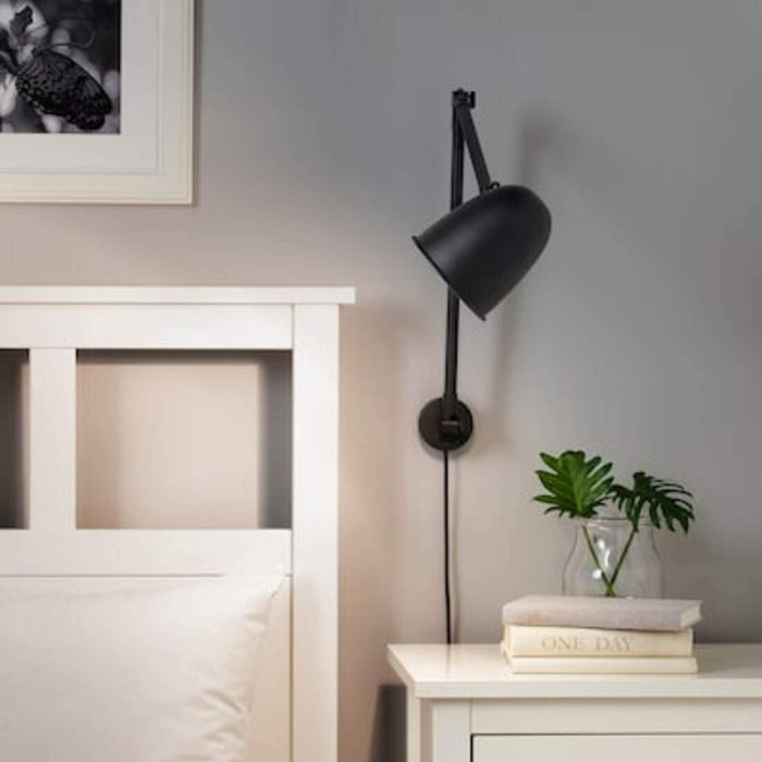 An IKEA black metal lamp with a cylindrical shade and a long adjustable arm, attached to a white wall.-40326025