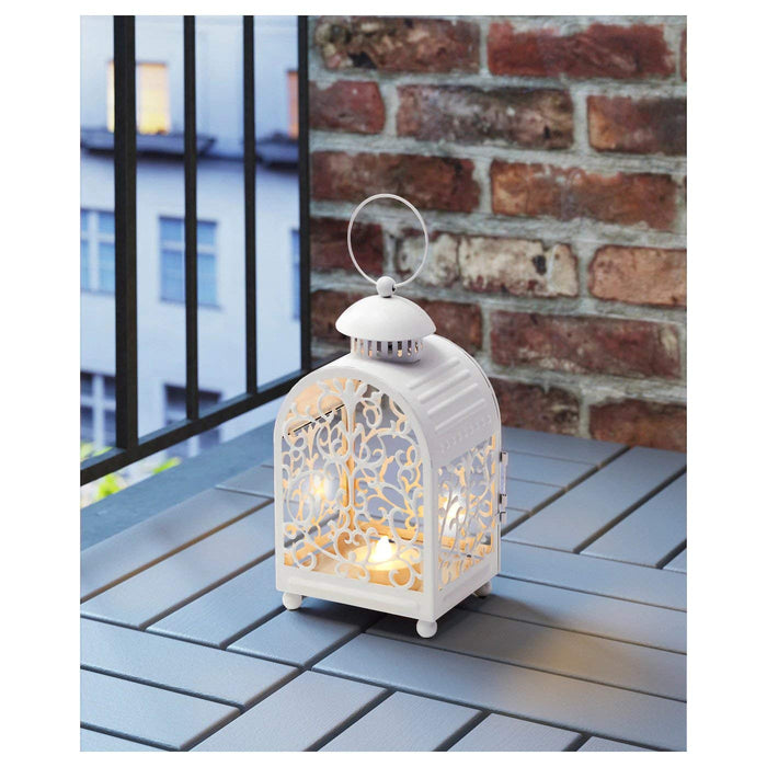 Image of the IKEA lantern with a metal cup and a lit candle inside, placed on a wooden table with a vase of flowers in the background. Alt text: "IKEA lantern with metal cup and candle on wooden table with flowers in background." digitalshoppy.in 20341475