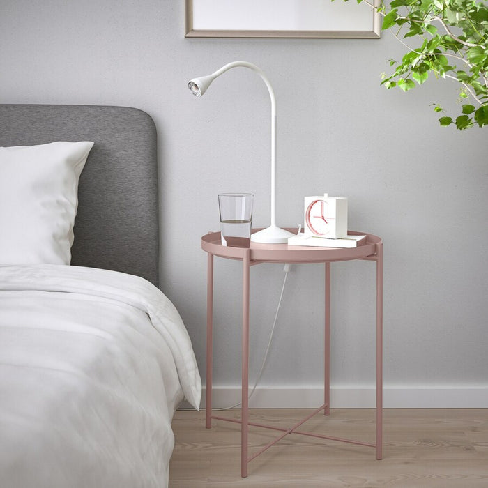 "The IKEA tray table in pale pink shown in a home setting. It is being used as a side table and has a vase of flowers and a book on top, highlighting its versatility."