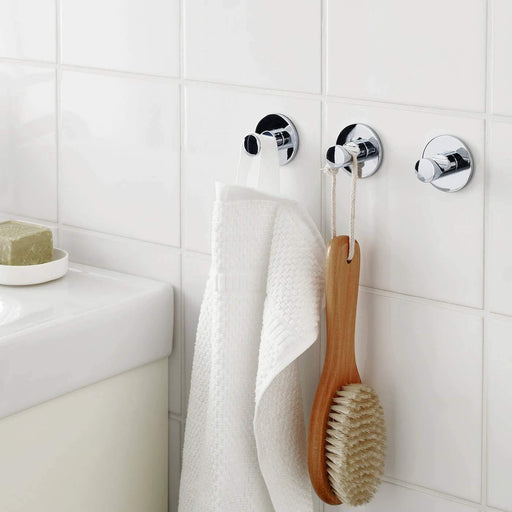 The IKEA Hook Self Adhesive - Chrome Plated, shown in use on a bathroom wall to hold a towel. The adhesive strip is visible on the back of the base