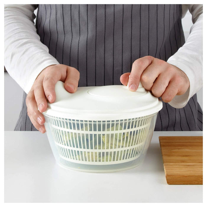 A hand holding a clear plastic IKEA Salad Spinner with lettuce leaves inside and water pouring over it. 30157235