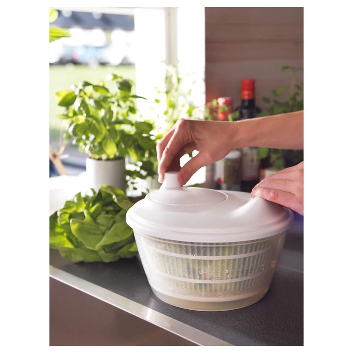 A person's hand holding the IKEA Salad Spinner while pouring water over a pile of mixed greens inside, A person's hand spinning the IKEA Salad Spinner's inner basket filled with lettuce, and water droplets flying out. 30157235